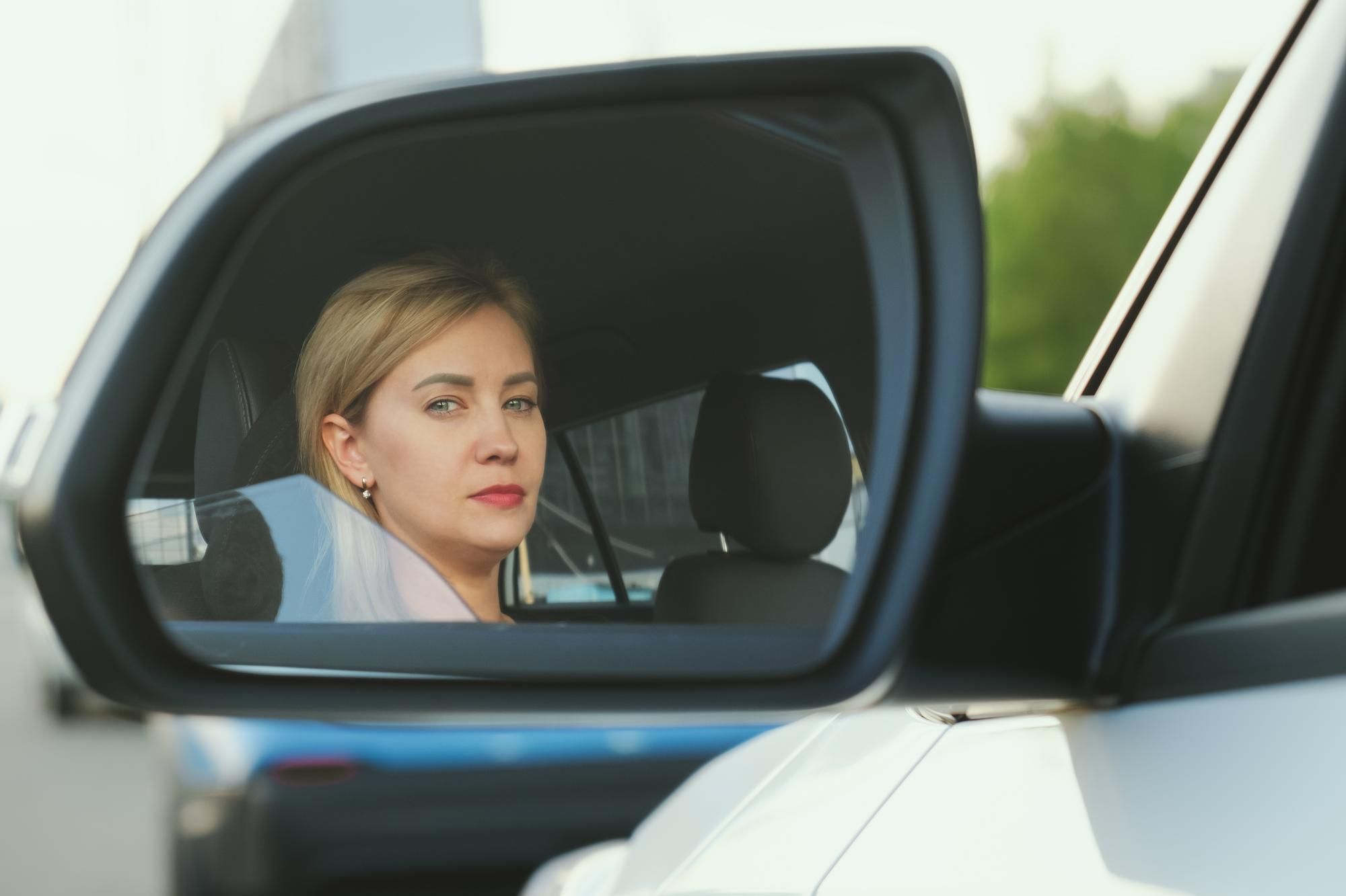 Una mujer reflejada en el espejo retrovisor lateral izquierdo.
