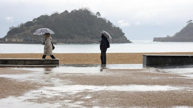 Dos personas pasean por la playa de la Concha durante un temporal de lluvia y frío.