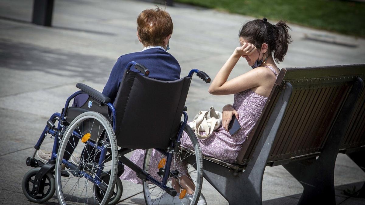 Una mujer mayor en silla de ruedas, acompañada de una cuidadora.