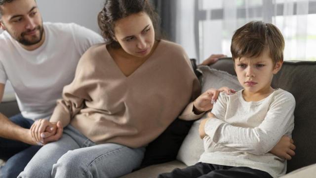 Un niño muestra enfado mientras permanece sentado en el sofá junto a sus padres.