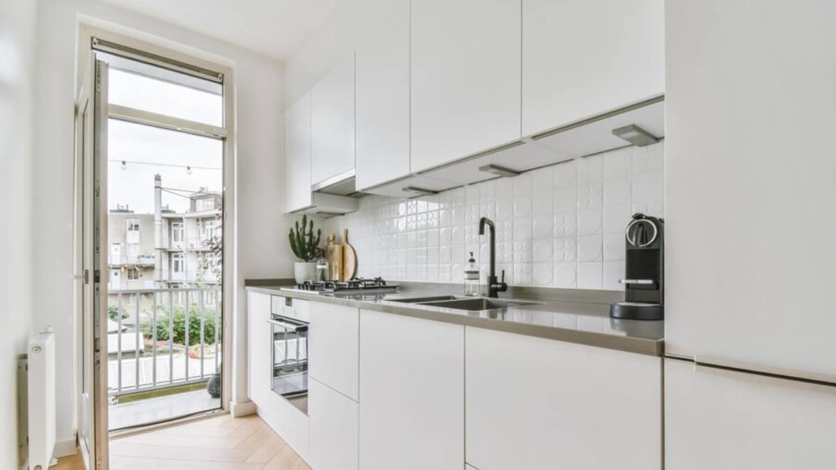 Cocina pequela en tonos blancos con la puerta del balcón abierta.