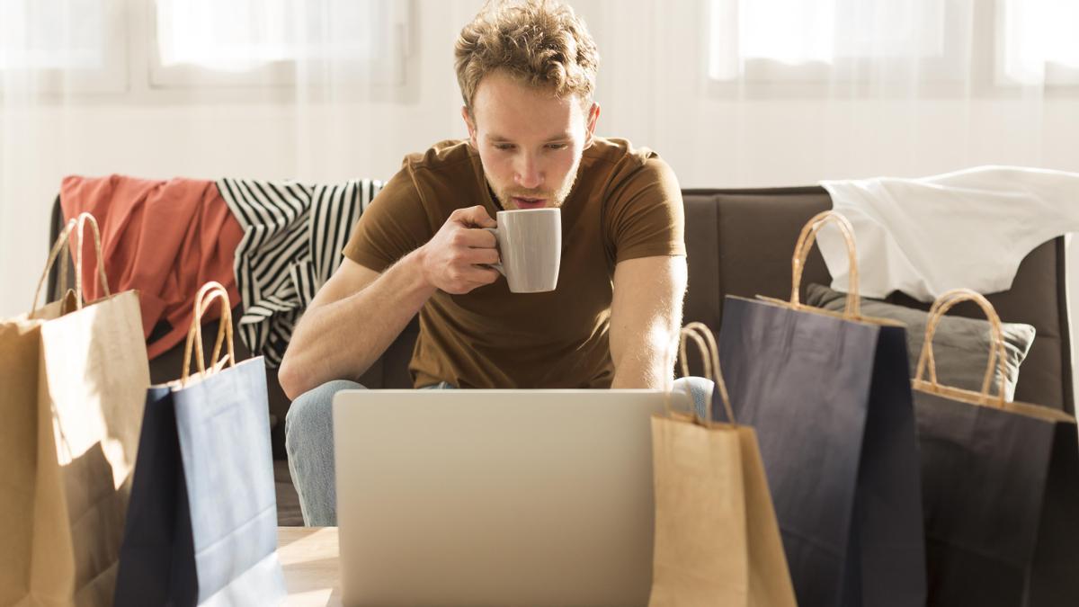 Hombre con el ordenador tomando café y rodeado de bolsas de compra.