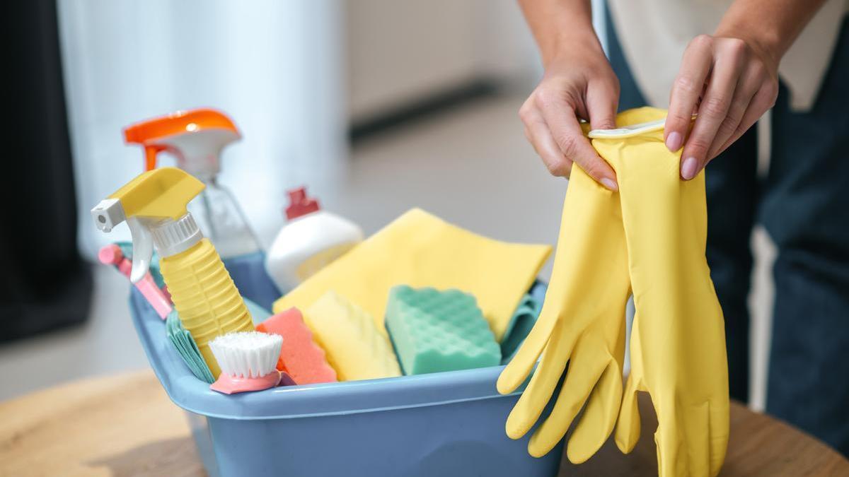 Una joven se pone unos guantes junto a un cubo con diferentes productos de limpieza.