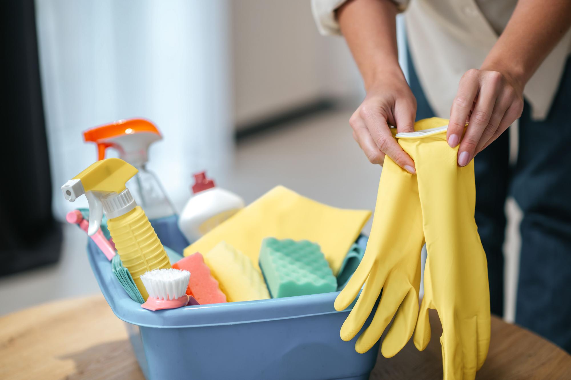 Chica se pone unos guantes junto a un cubo con diferentes productos de limpieza.