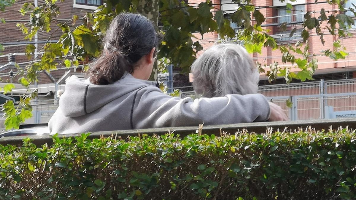 Una persona acompaña a una mujer mayor sentadas ambas en el banco de un parque.