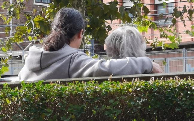 Una persona acompaña a una mujer mayor sentadas ambas en el banco de un parque. Una persona acompaña a una mujer mayor sentadas ambas en el banco de un parque.