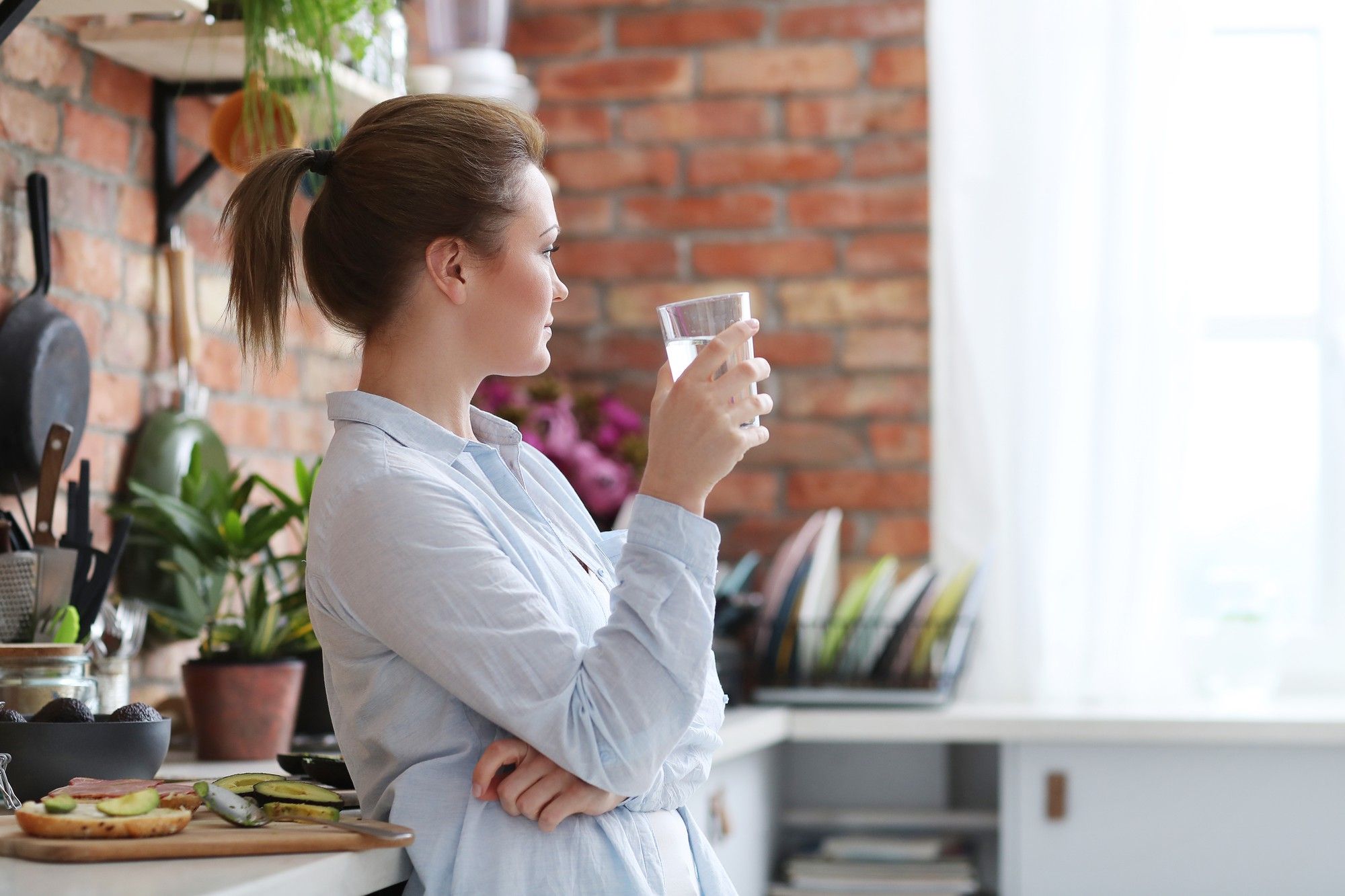 Una mujer bebe un vaso de agua mientras prepara el desayuno.