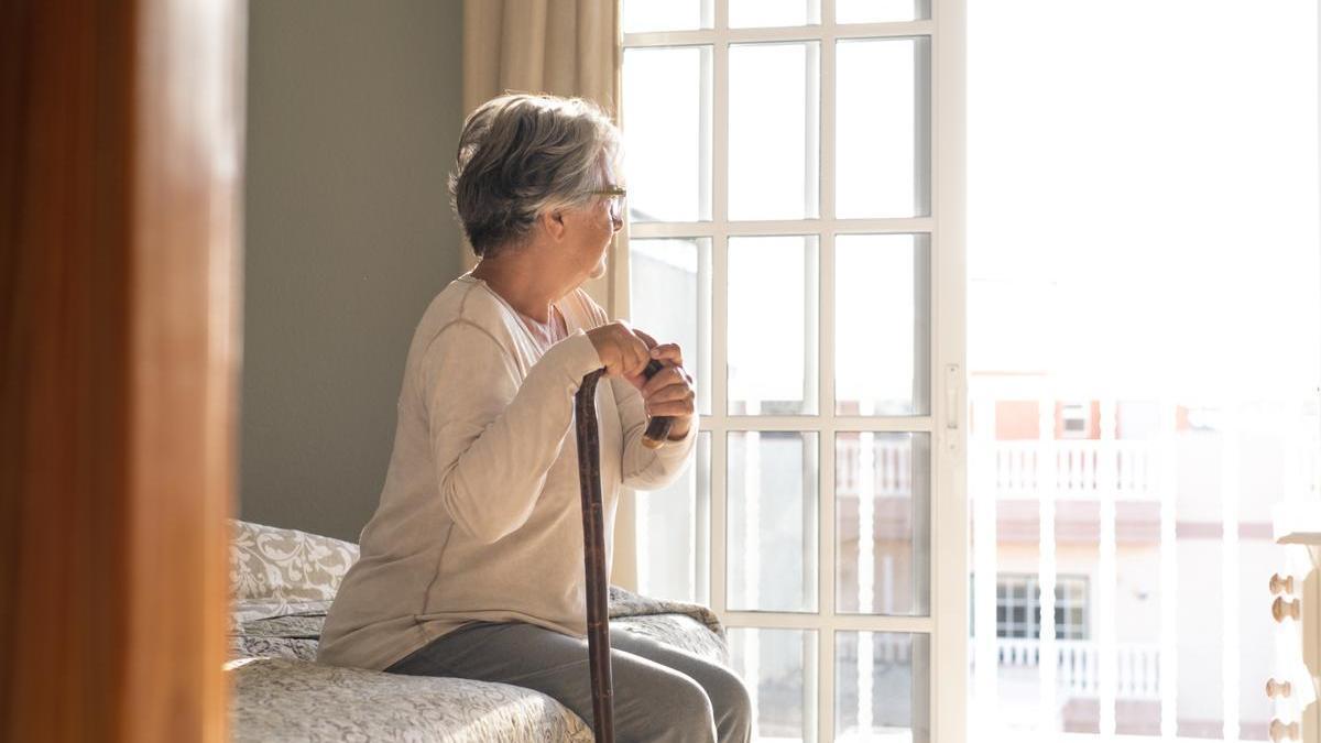 Señora mayor mira por la ventana sentada en el sofá de casa con un bastón en la mano.