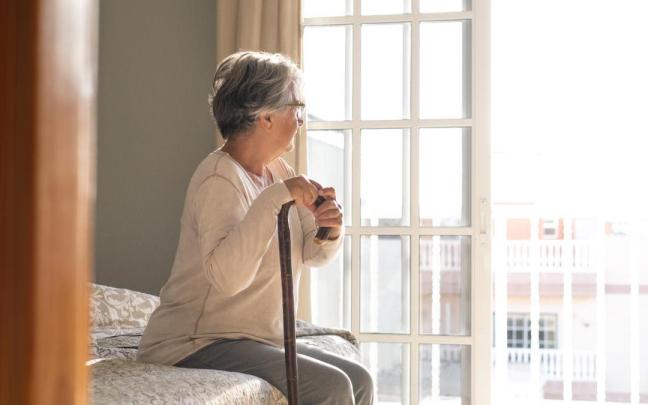 Señora mayor mira por la ventana sentada en el sofá de casa con un bastón en la mano.