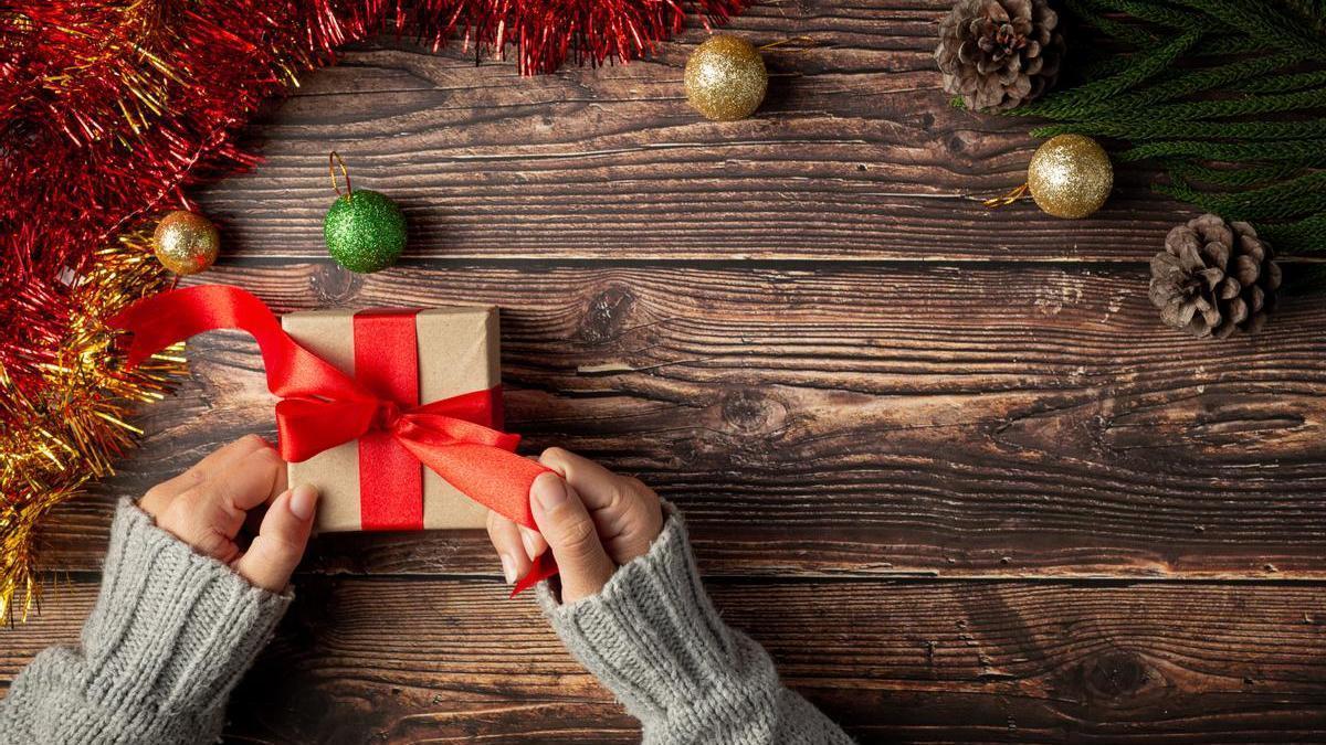 Manos envuelven un regalo y colocan un lazo de color rojo con decoración navideña de fondo