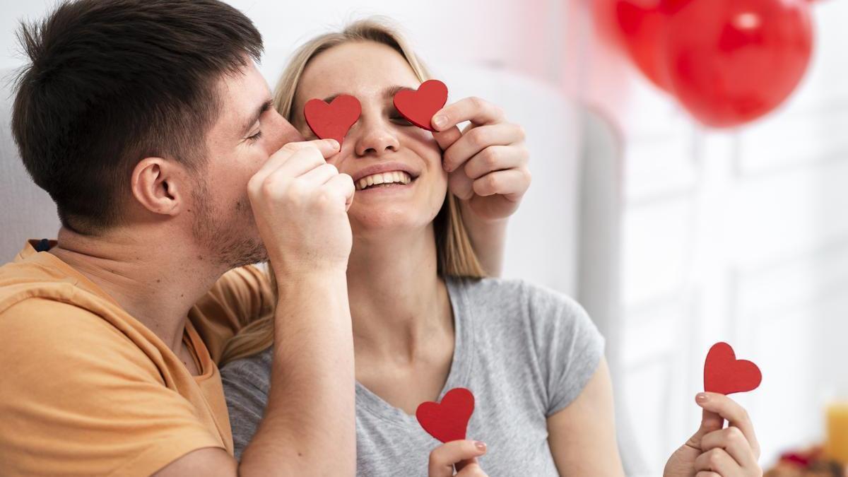 Pareja sonriente celebra el Día de San Valentín con globos y corazones de cartulina.