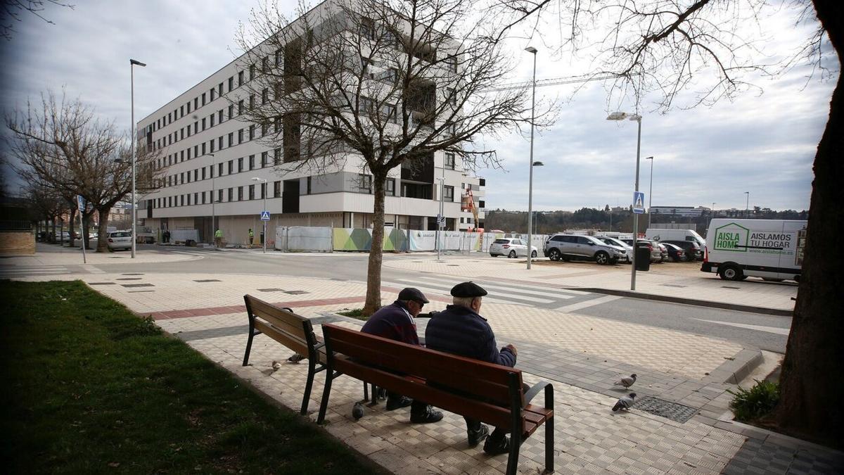 Dos personas mayores, en un banco junto a un bloque de viviendas