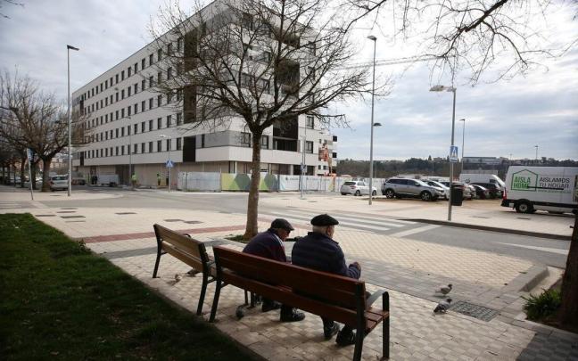 Dos personas mayores, en un banco junto a un bloque de viviendas