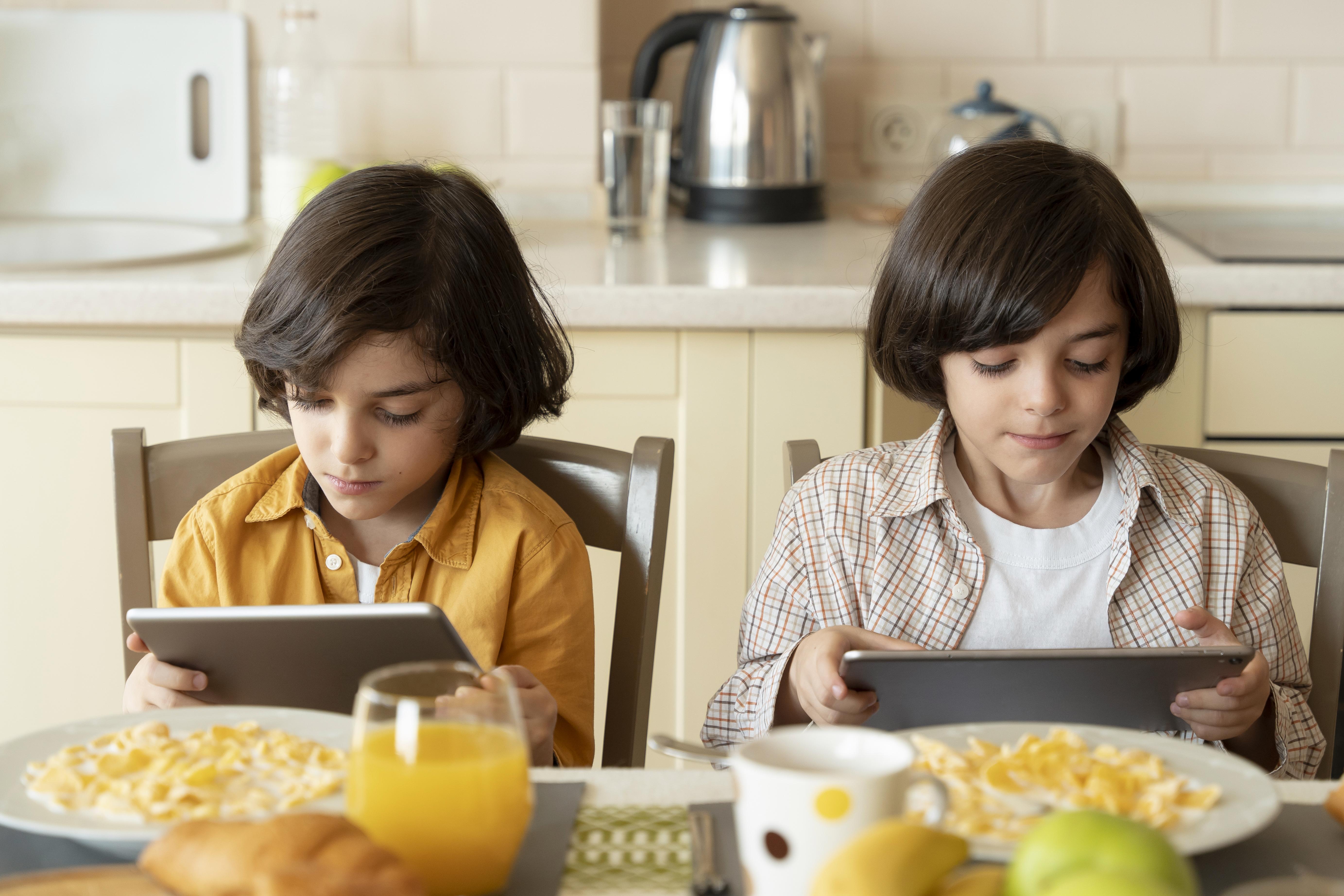 Dos niños comen mientras juegan con su tablet