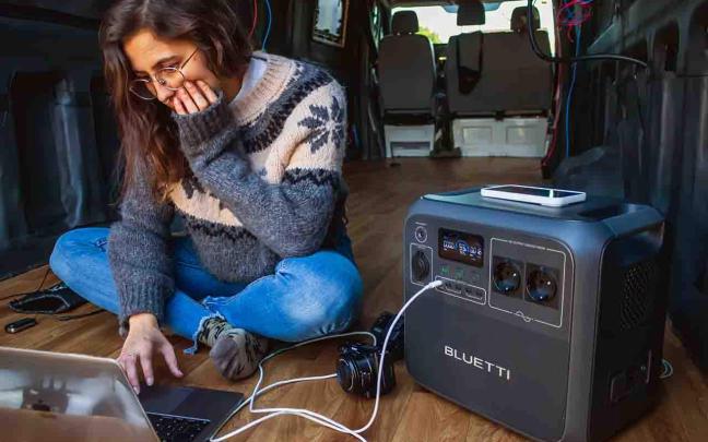 Una joven teletrabaja en su furgoneta utilizando una estación de energía portátil para cargar sus dispositivos